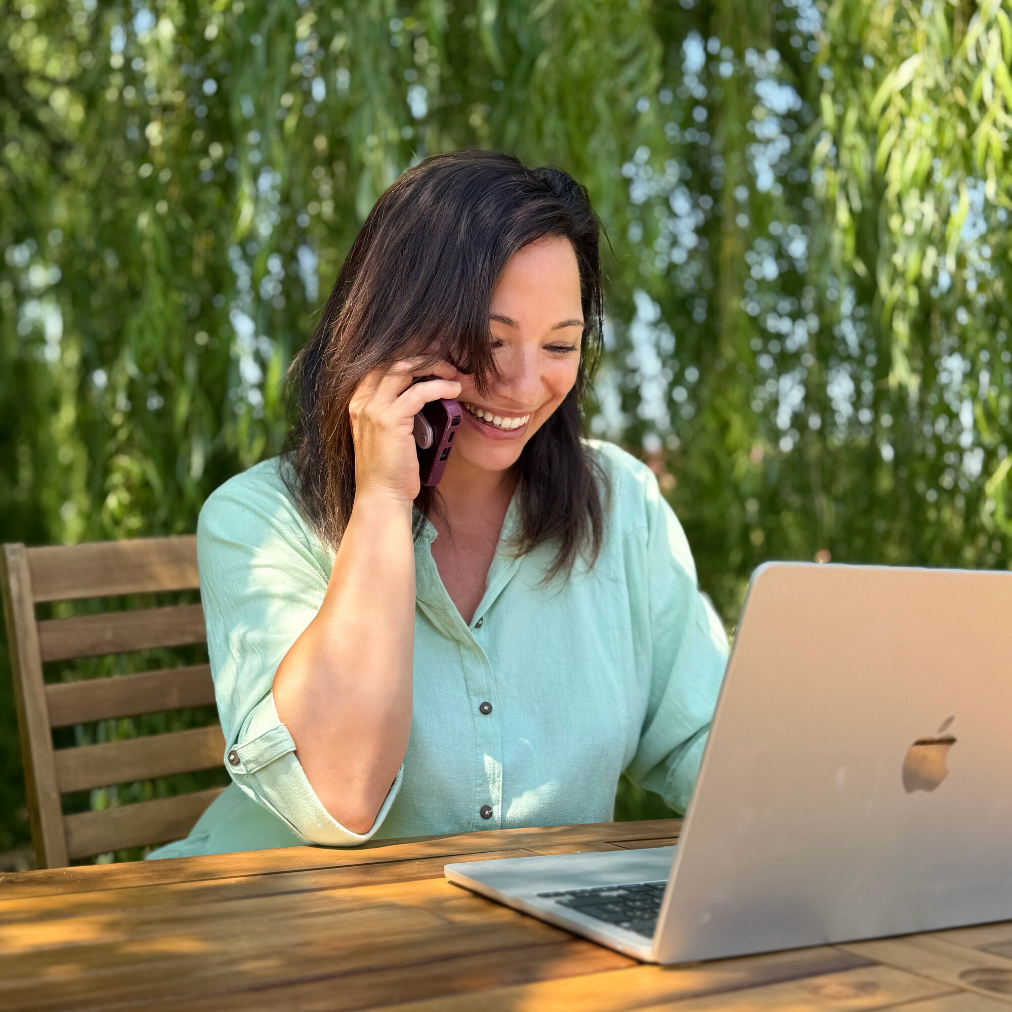 Silvia sorride parlando al telefono davanti al laptop, sotto le fronde di un salice